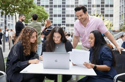 A group of four people, three women and one man, engaging with laptops and papers at a table outdoors.
