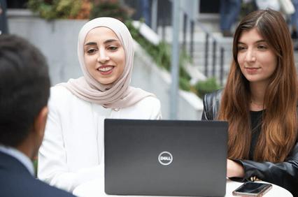 Two women, one wearing a hijab, are sitting around a table with a laptop, engaged in a conversation with a man whose back is to the camera.
