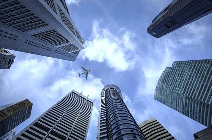 A plane flies above skyscrapers under a partly cloudy sky.