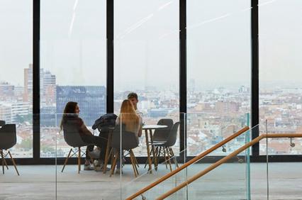 Three people are sitting at a table in a modern office with large windows overlooking the city.