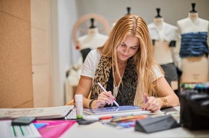 A young woman is sketching designs in a studio surrounded by dress forms.