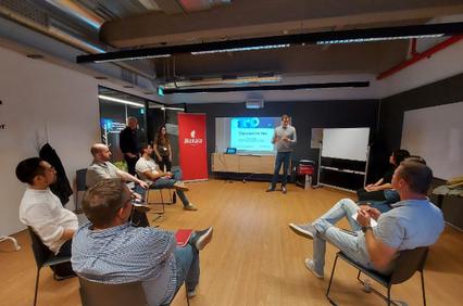 A man presenting in front of a small audience in a modern, informal setting, with a digital screen displaying a presentation in the background.