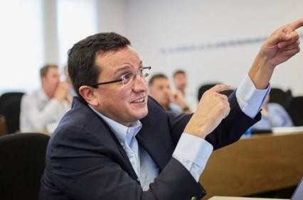 A man in a business suit is pointing and speaking during a meeting in a conference room.