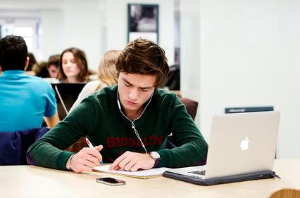 A young man studying in a busy university library while listening to music on his headphones.