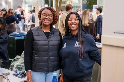 Two women smiling at an outdoor event, with other people in the background.
