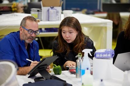 A man and a woman are collaborating at a table with various objects around them.