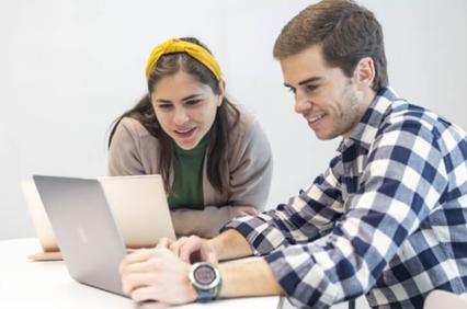 Two young adults, a male and a female, are looking at a laptop screen while sitting at a table.