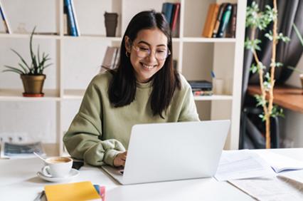 A young woman wearing glasses and a green sweater smiles while using a laptop at a desk with books and a cup of coffee.