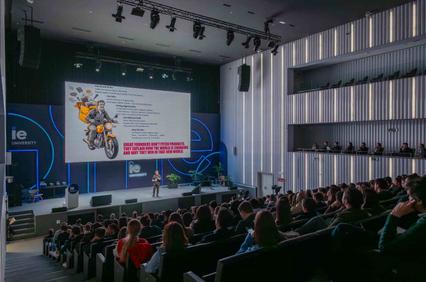 A speaker stands on stage in a modern auditorium with a large screen displaying a colorful presentation about entrepreneurship.