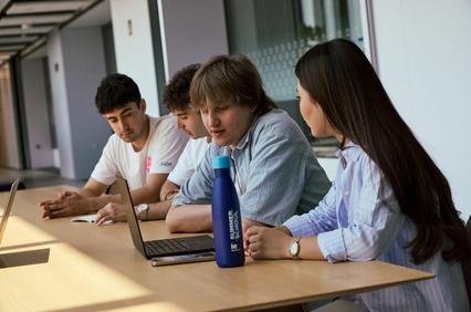 A group of four young people engaged in a discussion while working on a laptop at a table.