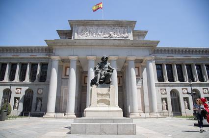 A view of the Prado Museum entrance featuring a statue in the foreground and the Spanish flag above.