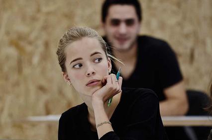 A young woman in a black top looks surprised while holding a phone to her ear in a classroom setting, with other students around her.