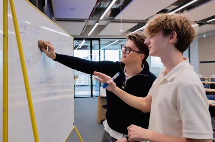 Two students are collaborating at a whiteboard in a classroom setting.