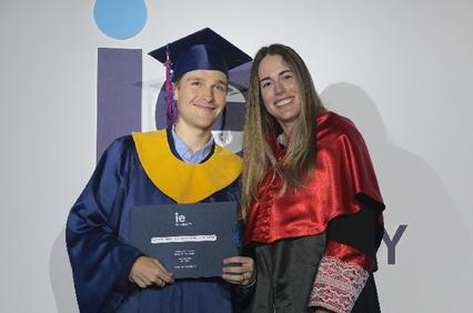 A young man in a blue and yellow graduation cap and gown holding a diploma, standing next to a young woman in a red graduation stole.