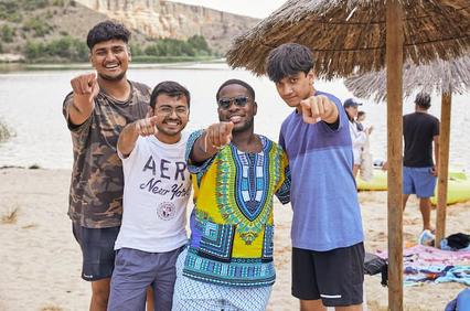 Four young men smiling and pointing towards the camera at a lakeside beach.