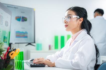 A laboratory scientist is working on a computer, analyzing data with green test tubes in the background.