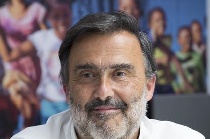 A middle-aged man with a beard is smiling in front of a blurred background featuring groups of people.