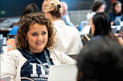 A smiling woman wearing a university shirt engages in conversation at a crowded workspace.