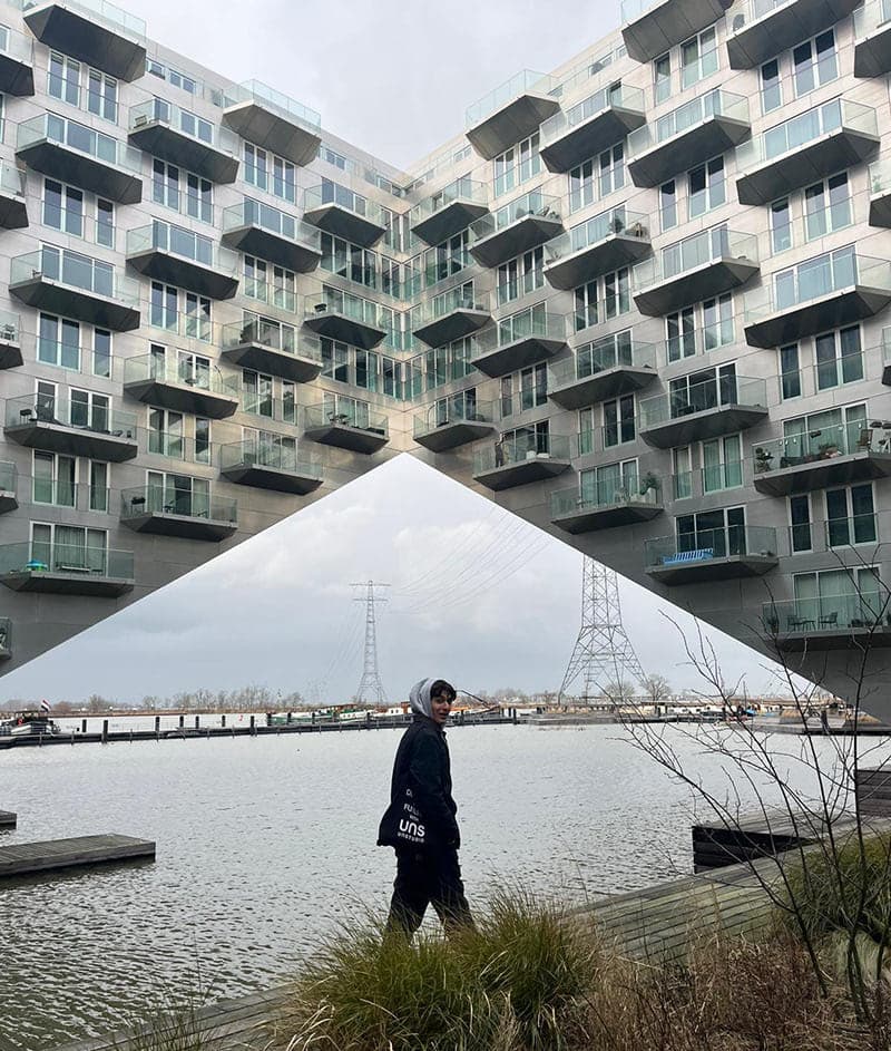 A person standing by a waterfront near a unique modern building with an architectural design featuring large balconies.