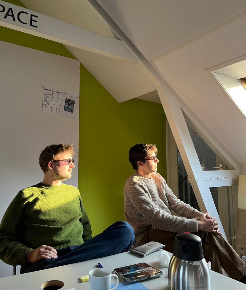 Two young men are sitting in a well-lit room with a green wall, deep in thought.