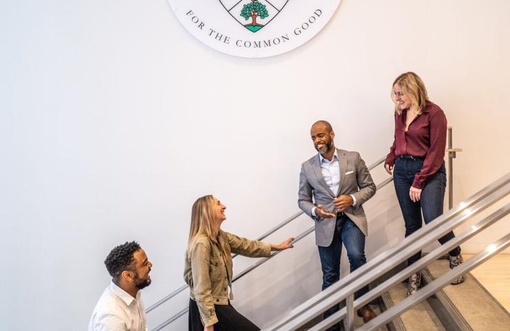 Four professionals are conversing on a staircase inside a building, beneath an emblem that includes the text 'Guildford College - For the Common Good'.