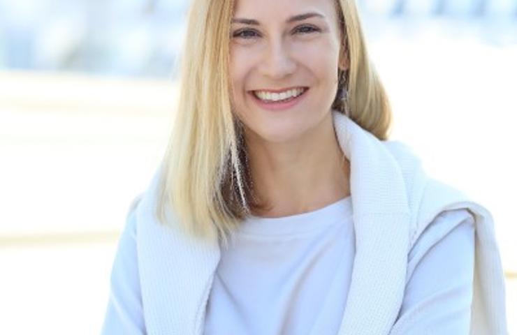 A smiling woman with blonde hair wearing a white top and a light sweater draped over her shoulders outdoors.