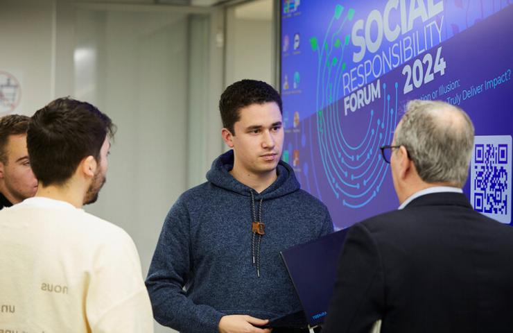 A young man engages in a conversation with an older man at a social responsibility forum, while others listen.