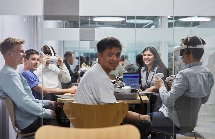 A group of young adults is engaged in a technology-focused meeting in a modern office, with one person using a virtual reality headset.