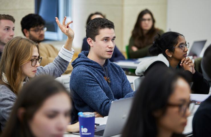 A young man raises his hand to ask a question in a university classroom filled with students.