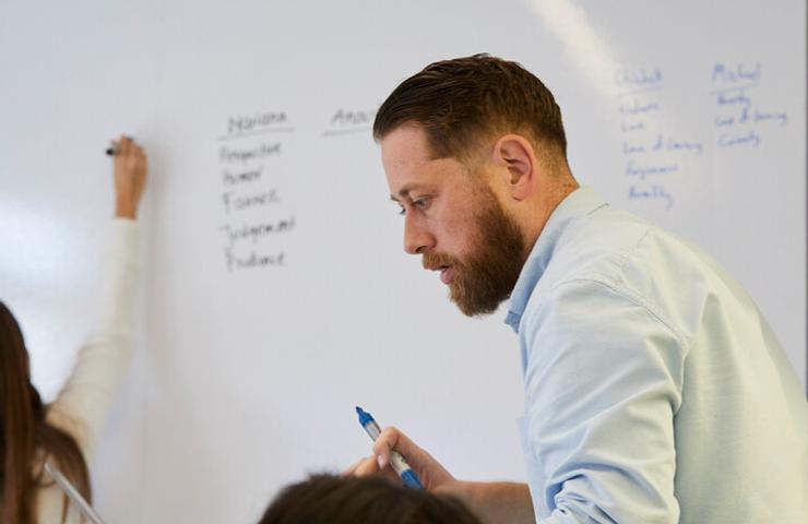 A business meeting with a man writing on a whiteboard while colleagues write notes.