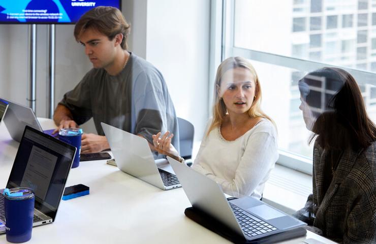 Three young adults are sitting at a table working on laptops in a modern office environment, engaged in a discussion.