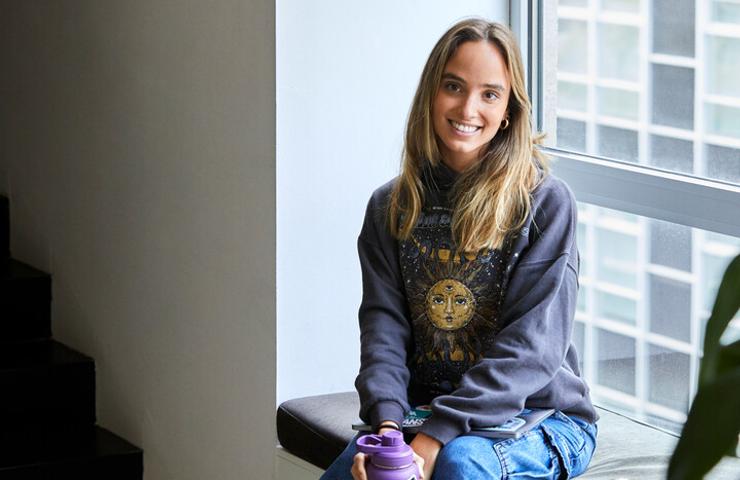 A young woman sits smiling in a casual setting, holding a purple bottle.