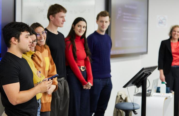 A group of young adults stands in a classroom, attentively watching a presentation.