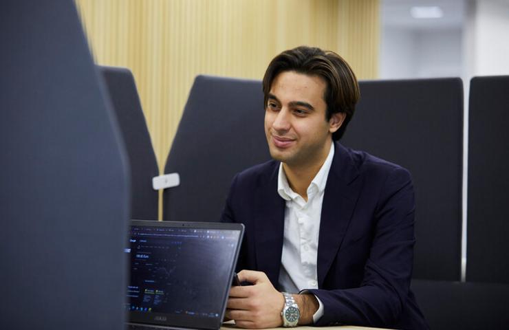 A man in a business suit smiling while working on a laptop in an office environment.