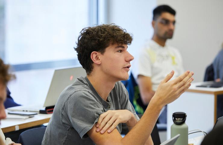 A young male student, engaging in a discussion in a classroom setting, with other students around him.