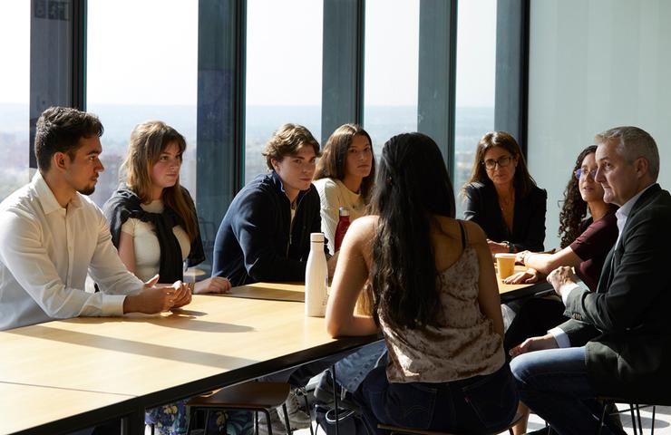 A group of seven people, including young adults and an older man, having a meeting around a table in a room with large windows.