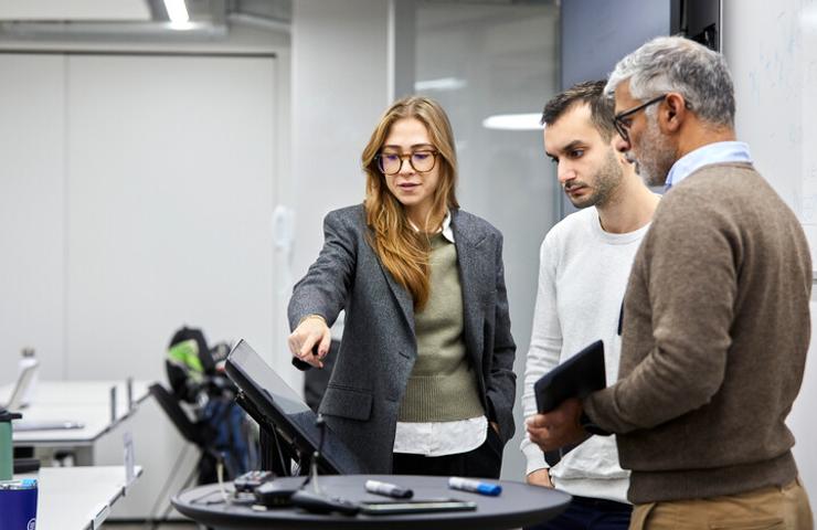 A woman gestures at a computer screen while discussing with two male colleagues in an office setting.