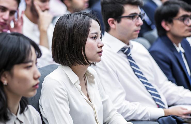 A group of young adults attentively listening in a seminar or class, predominantly wearing business attire.