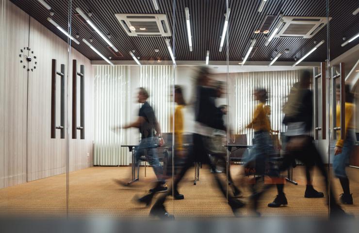 Blurry motion of busy people walking in a modern office corridor with wooden decor and glass partitions.