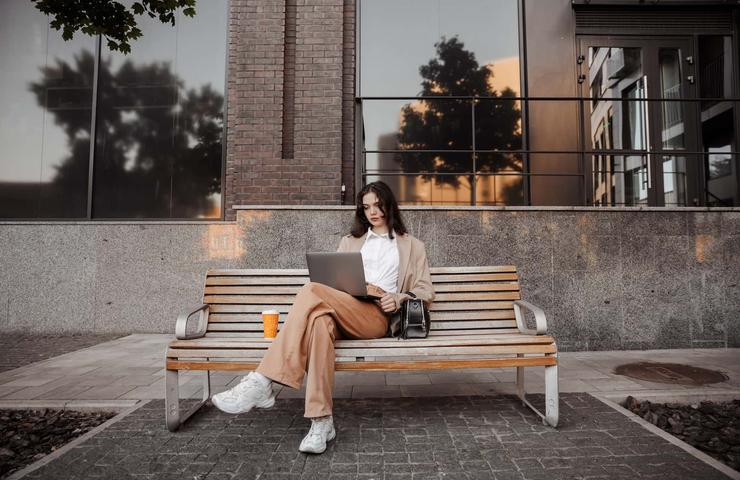 A woman sits on a bench outside a modern building, working on her laptop.