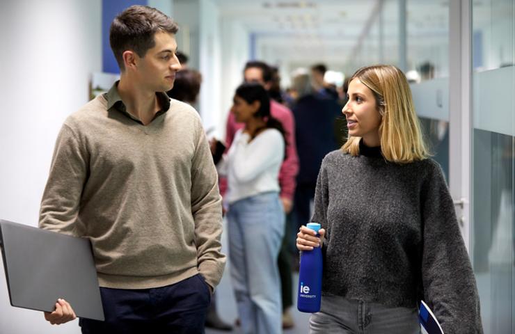 A man and a woman walking and talking in a busy office corridor, both carrying items; the man a laptop and the woman a water bottle.