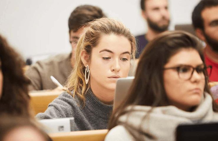 A focused young woman using a laptop in a classroom full of students.