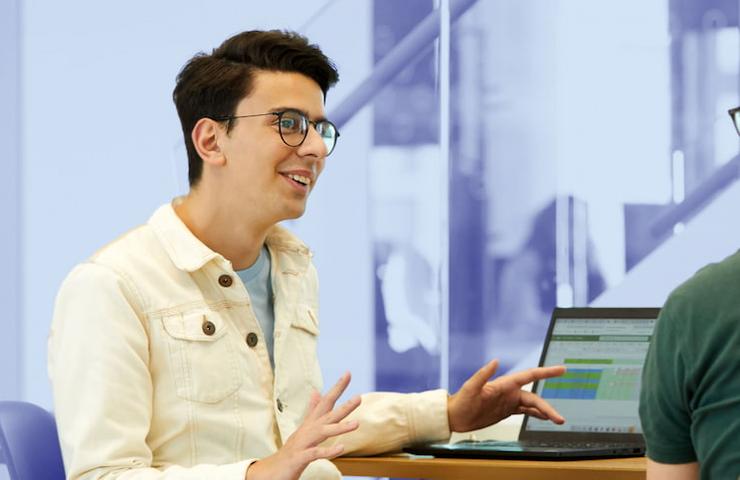 A young man wearing glasses and a light beige jacket is talking to another person at a table with a laptop.