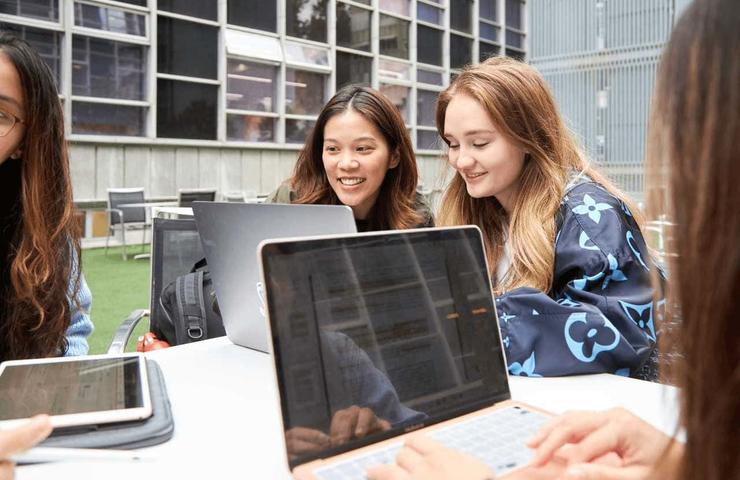 Three young women studying together with laptops and a tablet at an outdoor campus setting.