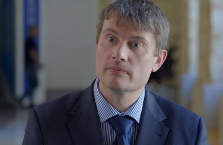 A man in a business suit with a concerned expression sits in an indoor setting.