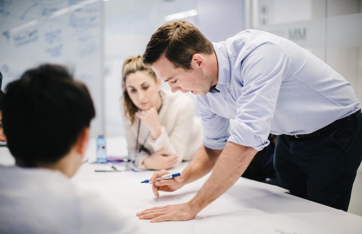 A man in a blue shirt is leaning over a table during a discussion with colleagues in an office setting.