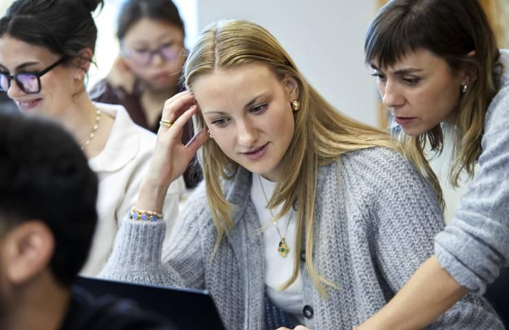 A woman in a gray sweater is looking at a computer screen while speaking on a mobile phone in an office environment, surrounded by other focused colleagues.