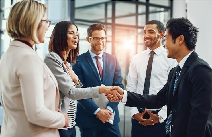 Two professionals shaking hands in a group of smiling colleagues in a modern office setting.