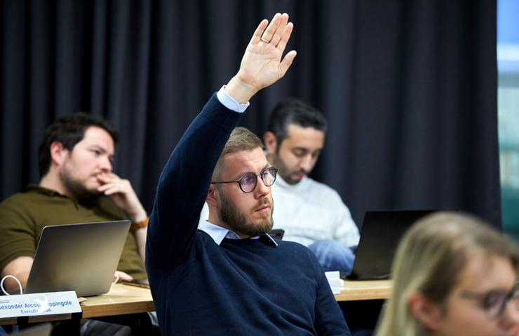 A man in a classroom setting raising his hand to ask a question or provide a response.