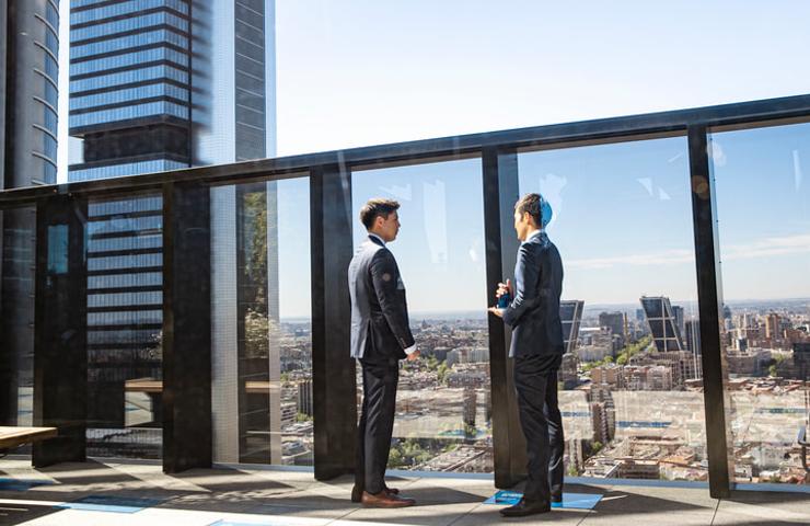 Two businessmen in suits having a discussion in a modern office building with a cityscape in the background.